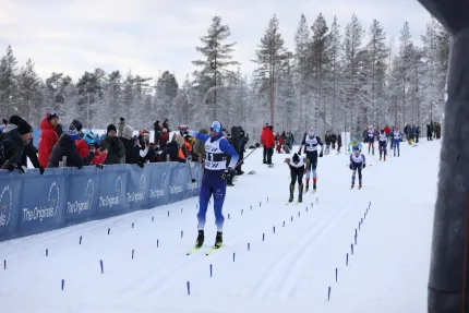 Längdskidåkare i blå dräkt glider mot mål medan publik står bakom avspärrning vid snöig skogsbana..
