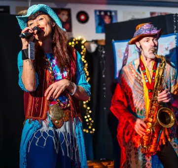 En kvinna sjunger i mikrofon bredvid en man som spelar saxofon, båda i färgstarka kläder inomhus..