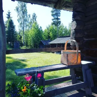Flätad korg med handtag står på en träbänk i en timmerbyggnad med grön gräsmatta och träd utanför..