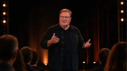 En man med glasögon står på en mörk scen och gestikulerar medan publikens huvuden syns i förgrunden..