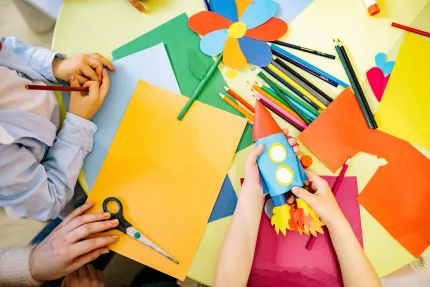 Barnhänder pysslar vid ett bord med färgat papper, sax, färgpennor och en pappersraket samt blomma..