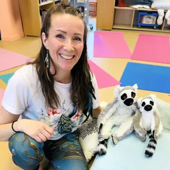 Leende kvinna sitter på golvet i ett rum med färgglada mattor och två lemur-mjukisdjur på en kudde..