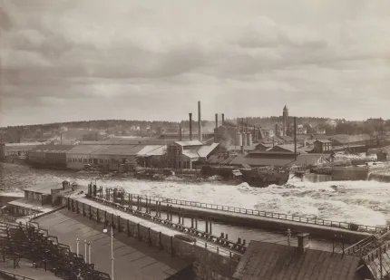 Svartvit vy över ett industriområde med fabriksbyggnader, skorstenar och forsande vatten vid en damm..