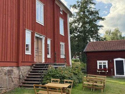 Rött trähus med vita fönster och trappa, samt en mindre röd byggnad bredvid, utemöbler och gräsmatta..