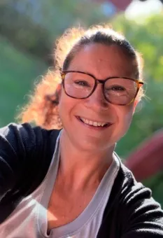 Närbild på en leende person med glasögon som tar en selfie utomhus i starkt solljus, med grön bakgrund..