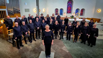 En grupp körsångare klädda i svart står i en kyrka; en kvinna står längst fram med orgel och glasmålningar i bakgrunden..