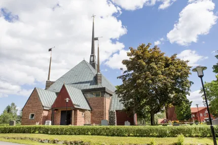 Röd tegelkyrka med grått plåttak och hög spira, träd och gravstenar under blå himmel med moln..