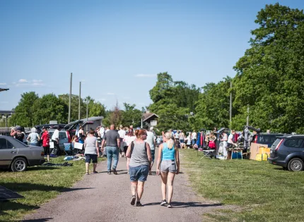 Folk promenerar längs en grusväg på en utomhusloppis med bilar, bord och klädställningar på gräs..