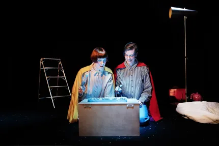 Två personer knäböjer vid en låda med blått ljus i mörk studio, med stege, lampa och säng i bakgrunden..