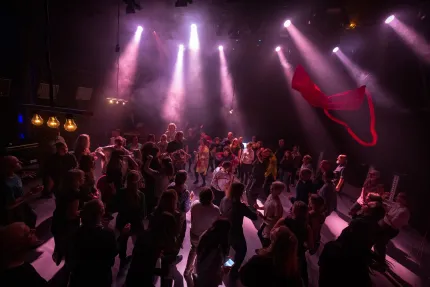 Folkmassa som dansar på ett dansgolv under lila scenbelysning och rök, med ett rött tyg som svävar i luften..