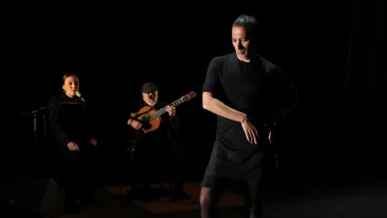 En flamencodansare i svart står i förgrunden, med sångerska och gitarrist i bakgrunden på mörk scen..