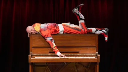 En man i röd mönstrad overall ligger tvärs över ett brunt piano på en scen med mörkröd ridå i bakgrunden..