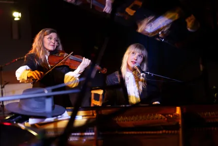 Två musiker på scen: en spelar violin och en sjunger i mikrofon vid ett piano, belysta av scenljus..