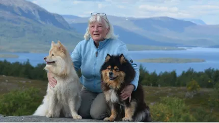 En äldre kvinna i blå jacka sitter utomhus med två hundar framför fjäll och en sjö i bakgrunden..