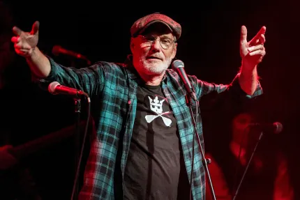 Äldre man på konsertscen med keps och glasögon står vid mikrofon med armarna utsträckta i rött scenljus.