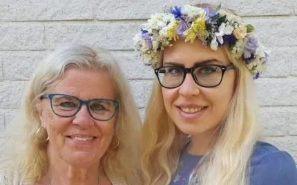 Närbild på två leende kvinnor med glasögon framför en ljus tegelvägg; den ena bär en blomkrans..