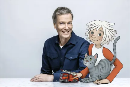 En leende man i blå skjorta sitter vid ett bord bredvid en tecknad figur med katt och leksaksbrandbil..
