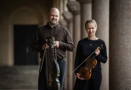 Två musiker står i en pelargång; en håller en nyckelharpa och den andra en violin med stråke..