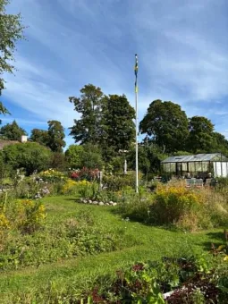 Blomstrande trädgård med gräsmatta, buskar och träd, en hög flaggstång samt ett växthus under blå himmel..