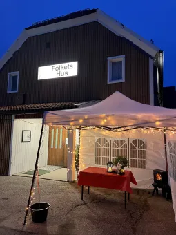 Byggnaden Folkets Hus i skymning med upplyst skylt, tält med ljusslingor och bord med dekoration utanför..