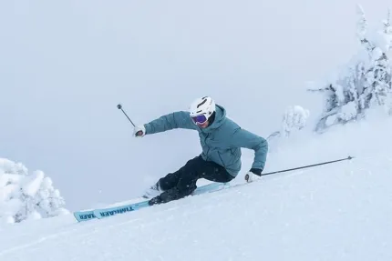 En skidåkare i hjälm och goggles svänger nedför en brant, snötäckt sluttning med snöiga träd i bakgrunden..