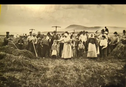 Sepiafärgat gruppfoto av bönder och familjer på en åker med liar och räfsor, med berg i bakgrunden..
