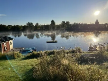 Still sjö i morgonsol med dimslöjor, brygga och roddbåtar vid strand, trädlinje speglas i vattnet..