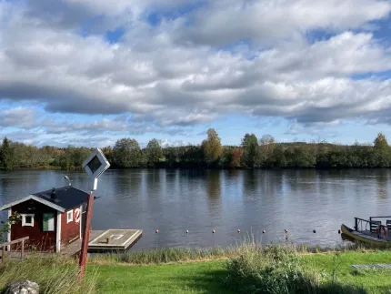Utsikt över en bred flod med molnig himmel, grön strand, liten bod och brygga samt båt vid kanten..