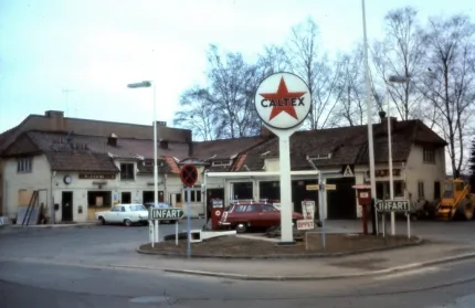 Gatumiljö med äldre bensinstation och rund Caltex-skylt, bilar vid pumparna och träd utan löv i bakgrunden..