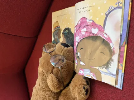 En brun teddybjörn med glasögon ligger i en röd soffa bredvid en uppslagen bilderbok med illustration..