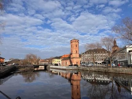 Rött tegeltorn vid en kanal i stadsmiljö, med spegling i lugnt vatten under blå himmel med moln.