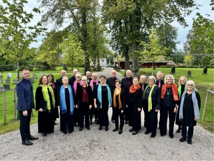 En grupp människor i svarta kläder med färgglada sjalar står på en grusgång i en grön kyrkogård med träd..
