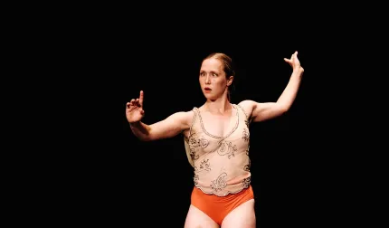 En dansare i beige linne och röda shorts poserar med utsträckta armar mot svart bakgrund på scen..