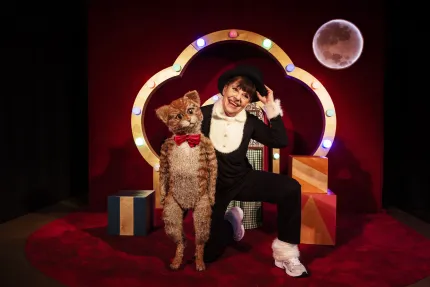 En leende scenartist i svart kostym och hatt poserar bredvid en kattfigur med rosett, framför ljusbåge..