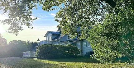 Ett ljust hus med mörkt tak ligger delvis dolt bakom grönska, med gräsmatta och sol lågt på himlen..