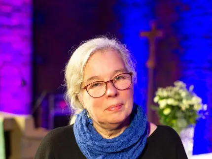 Närbild på en kvinna med glasögon och blå stickad sjal, framför en bakgrund i blått och lila ljus..