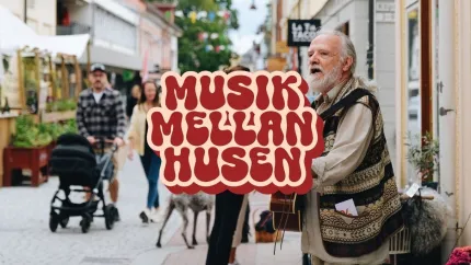 Äldre man spelar gitarr och sjunger på en gågata; förbipasserande och barnvagn syns i bakgrunden..