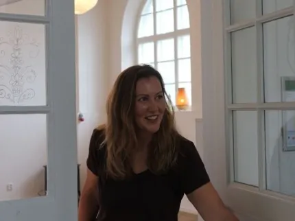 En leende kvinna står i en dörröppning i ett ljust rum med stora spröjsade fönster och vita väggar..