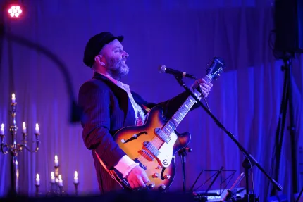 En leende musiker i hatt spelar elgitarr vid mikrofon på scen med blått ljus och tända ljus i bakgrunden.
