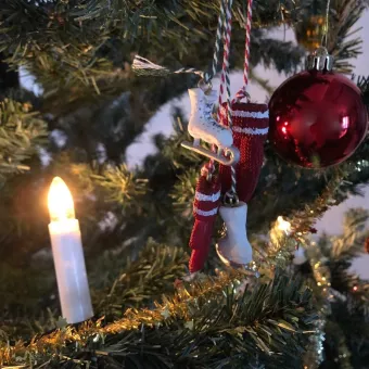 Närbild av julgran med röd julkula, hängande skridskoprydnad och tänd ljusliknande lampa bland grenar..