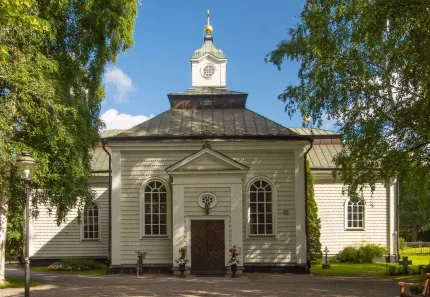 Frontal vy av en vit kyrkobyggnad med mörkt tak och klocktorn, omgiven av gröna träd under blå himmel..