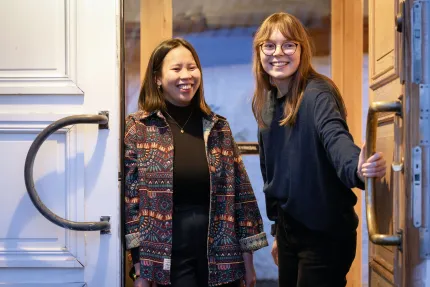 Två leende personer står i en öppen dörröppning inomhus, en håller i dörrhandtaget och tittar mot kameran..