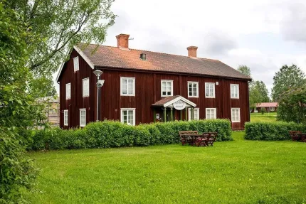 Stort rött trähus med tegeltak och vita fönster, omgivet av buskar och gräsmatta med utemöbler..