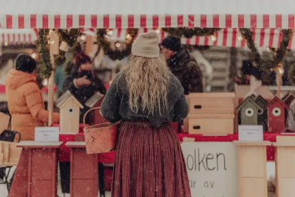 Person med grå mössa och långt hår står vid ett marknadsstånd med rödvit markis, trävaror och fallande snö..