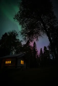 Mörk skog med siluetter av träd, en liten stuga med upplysta fönster och norrsken mot stjärnhimmel..