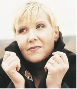 Närbild av en blond person som håller i kragen på en mörk jacka och blickar åt sidan mot ljus bakgrund..