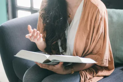 Person sitter i en soffa med en uppslagen bok i knät och gör en gest med handen i mjukt dagsljus..