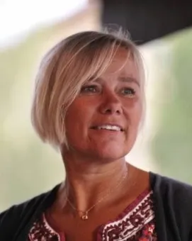 Närbildsporträtt av en blond kvinna med kort hår och halsband, iklädd kofta och mönstrad blus mot suddig bakgrund..
