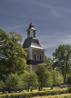 Kyrktorn med klocka och kors reser sig över lummiga träd vid en kyrkogård under klarblå himmel..