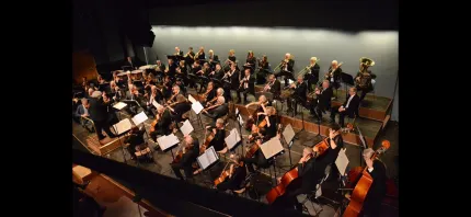 Ett symfoniorkester framträder på en scen med dirigent, stråkar och blåsare, noter på ställ och varmt ljus..
